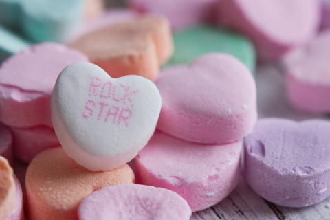 A mound of pastel candy hearts with printing on them on an aqua surface. One white heart has readable text: "Rock Star".