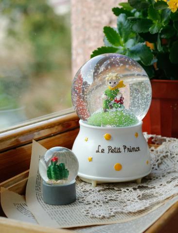 larger snow globe with a boy figurine and a smaller snow globe with a decorated tree on a tray next to a window