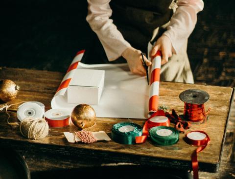 woman using scissors to cut gift wrapping paper. she is surrounded with ribbons, tape, and bows