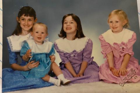 Image: Three girls and one baby sit on floor in matching dresses in front of grey-blue background Text: Olan Mills 89