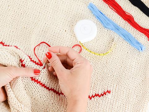A pair of hands creates a decorative stitch on cream fabric using red thread.