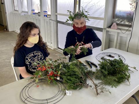Two adults sit at a table, adding fronds to empty wreath forms.