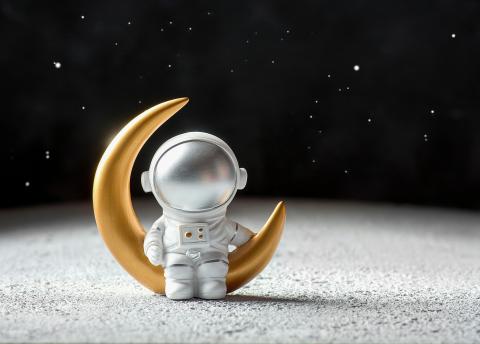 small silver astronaut sitting on a cold crescent moon on a white table and black background to simulate the moon