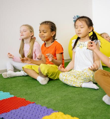 three children sitting on a green mat doing a cross-legged yoga pose with their eyes closed