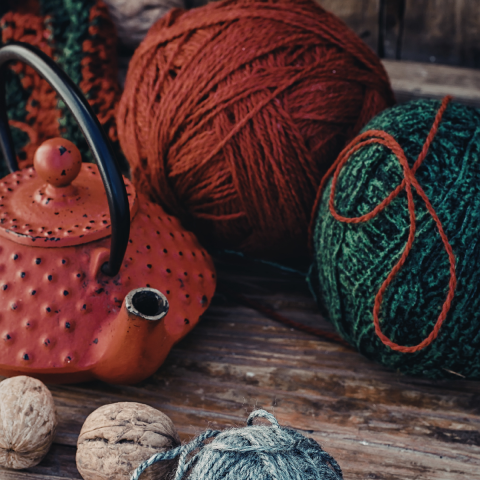 A close-up photo of a red teapot, several balls of yarn and some walnuts sitting on a wooden table