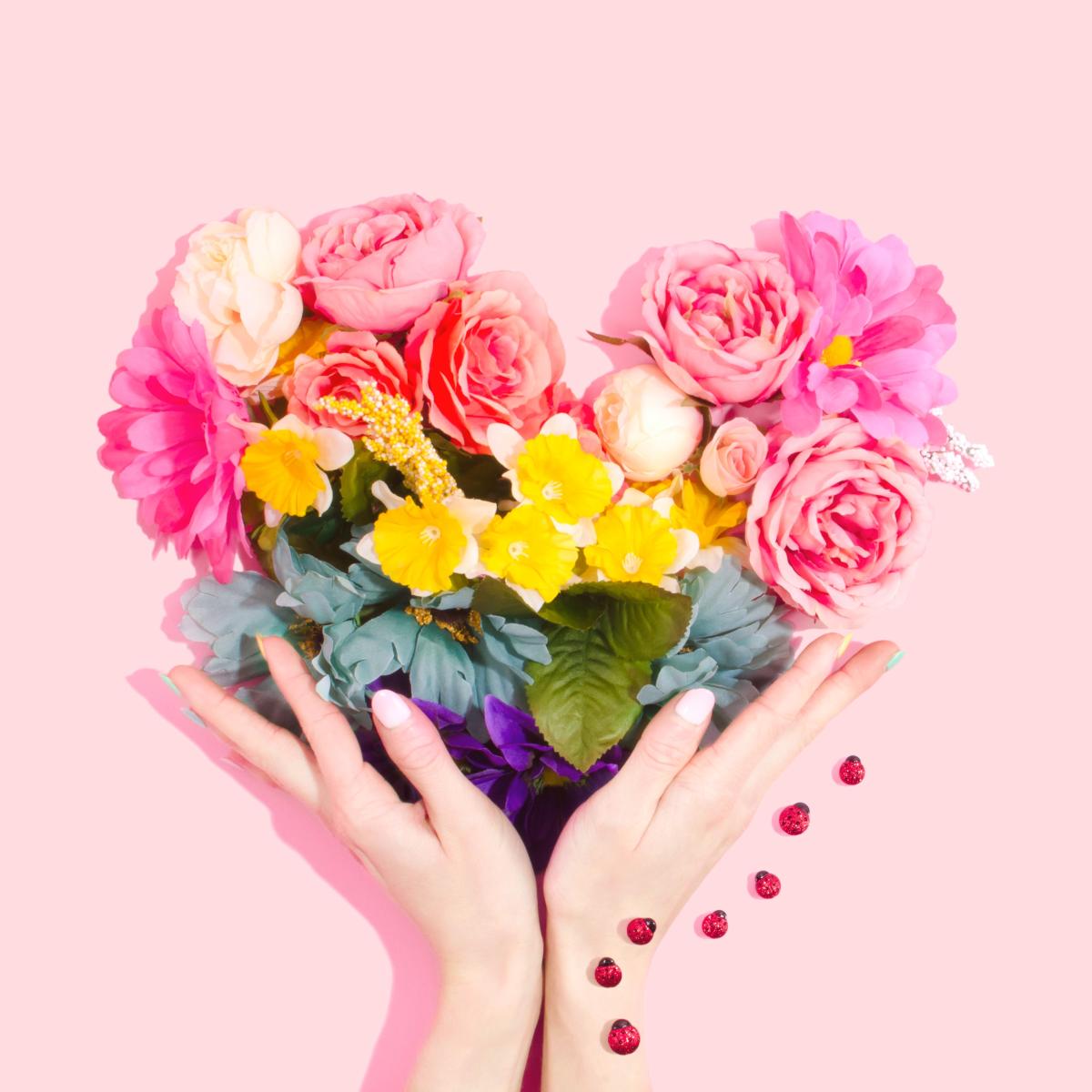 On a pink background, two hands offer up a heart made up of many different flowers.