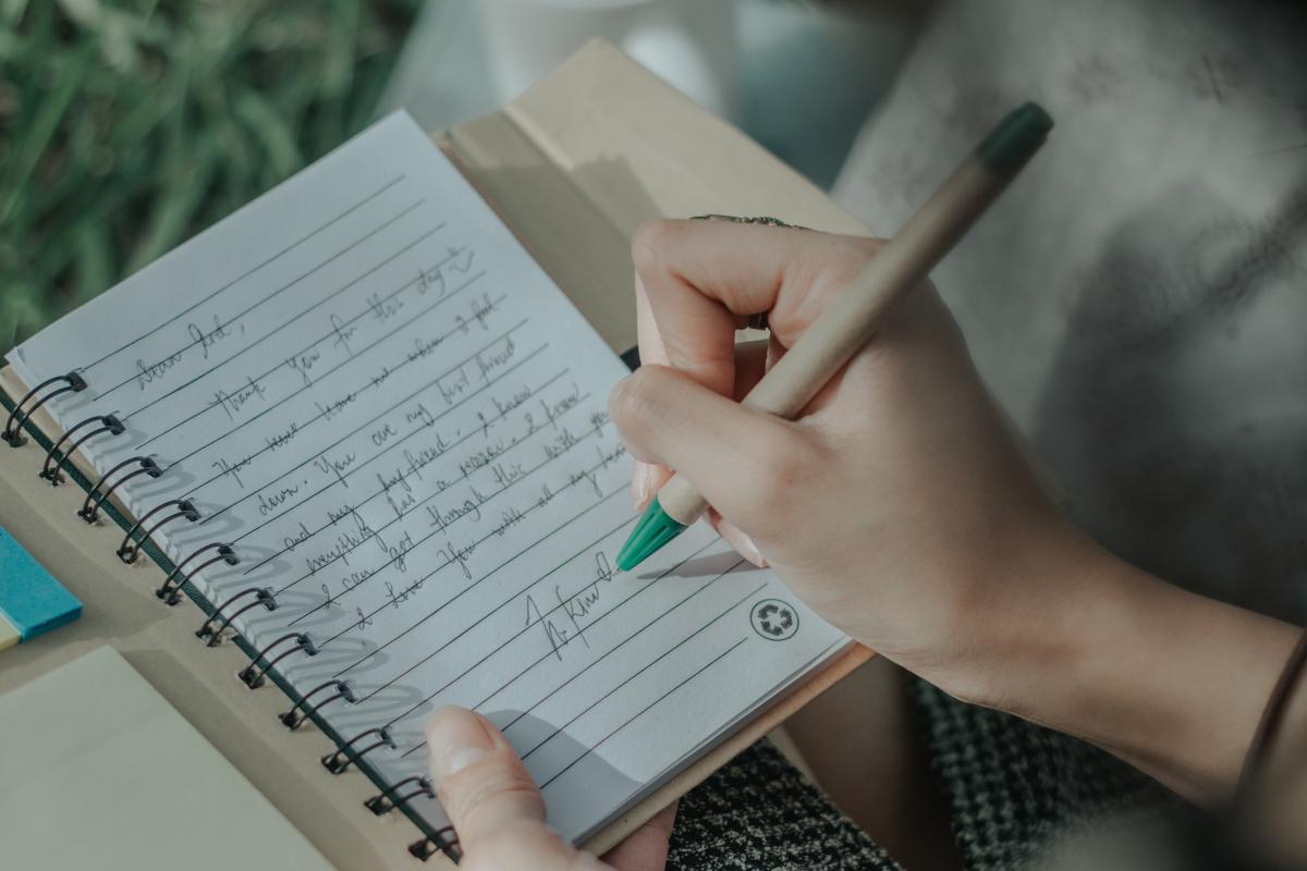 A person holds a spiral-bound journal in one hand and holds a pen in the other, writing on a half full page of handwritten text.