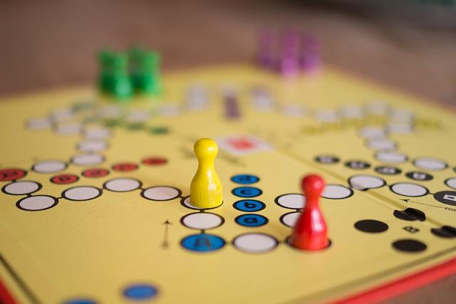 A yellow board game with white circle paths for game pieces to follow around the board. A yellow and a red pawn-shaped piece are in the foreground. Groups of four green and four purple pieces are blurry in the background.