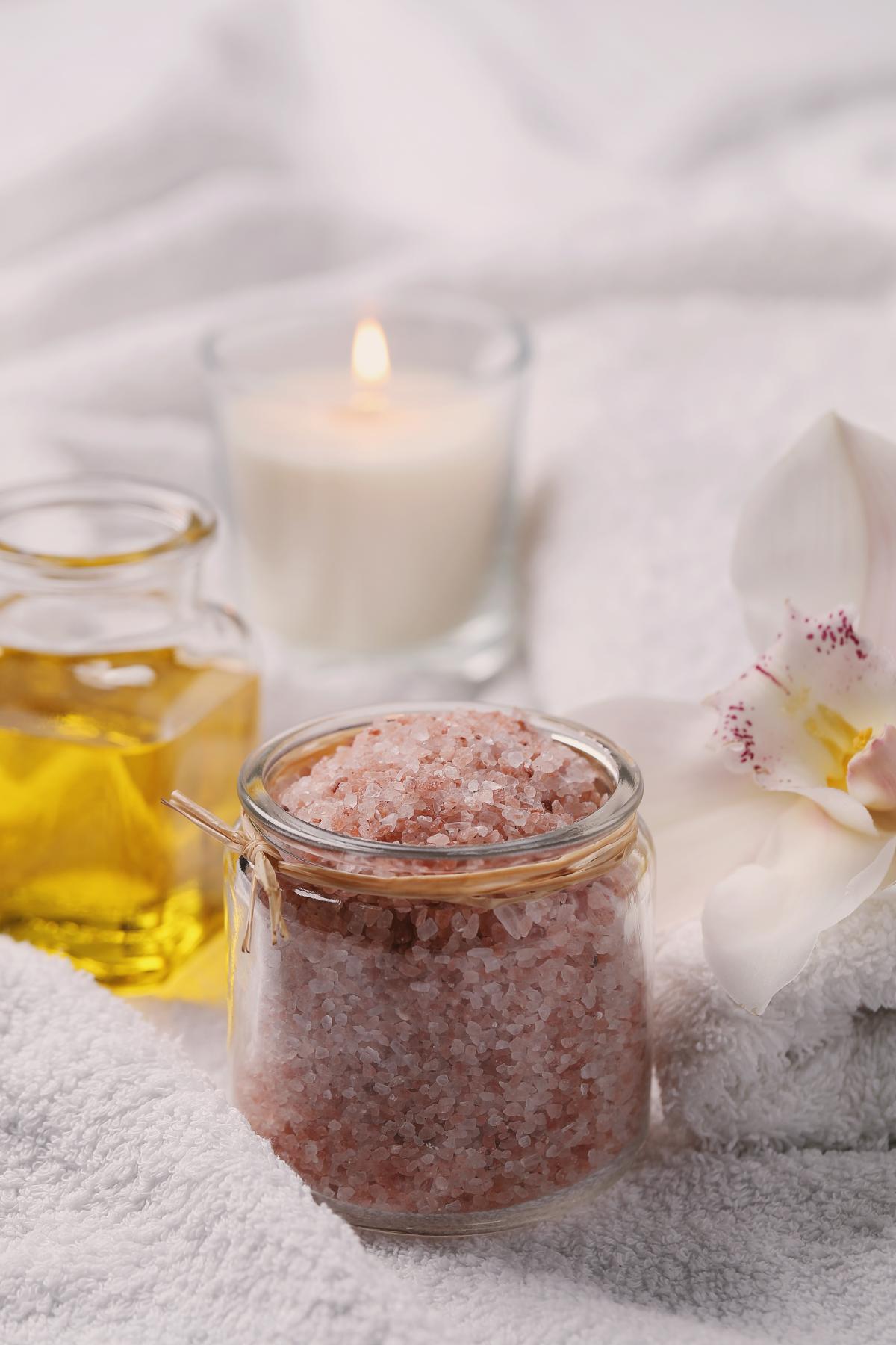 A small glass jar filled with pink salt scrub sits on a white towel. There is a small glass jar of oil behind it to the left, and a lit white candle in a glass jar directly behind the salt scrubs. There is a white lily to the right of the salt scrub.