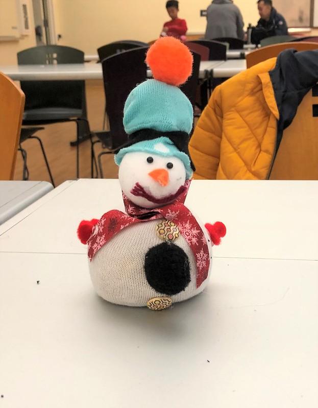 An example sock snowman made with a white sock body and blue sock hat. The snowman has buttons with geometric designs on them and a black pom pom on its stomach. A red ribbon with white snowflakes on it is used as a scarf. There is a large orange pom pom on its hat.