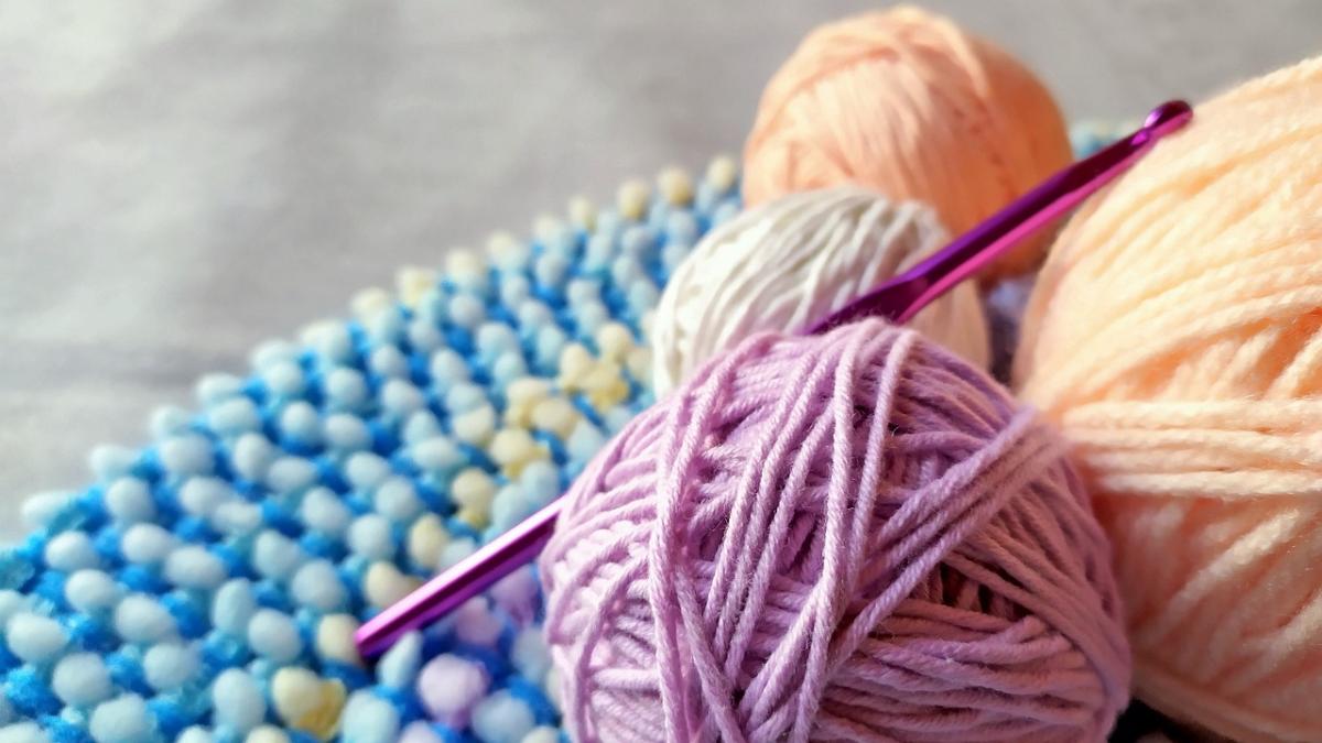 four yarn balls of different colors rest on a multicolored mat. A crochet needle rests in between two of the yarn balls. cuatro ovillos de diferentes colores descansan sobre una alfombra multicolor. Una aguja de crochet descansa entre dos de los ovillos de hilo.