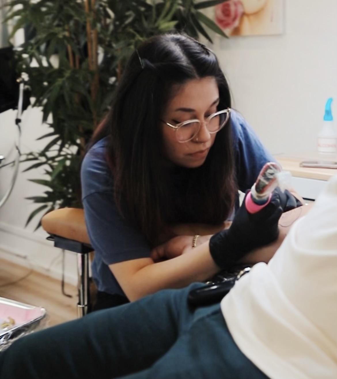 lady with dark hair wearing glasses tattooing a person out of sight but laying down