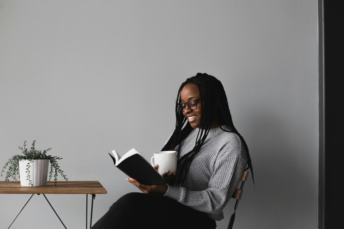 smiling young woman holding a warm beverage reading a book while sitting at a table