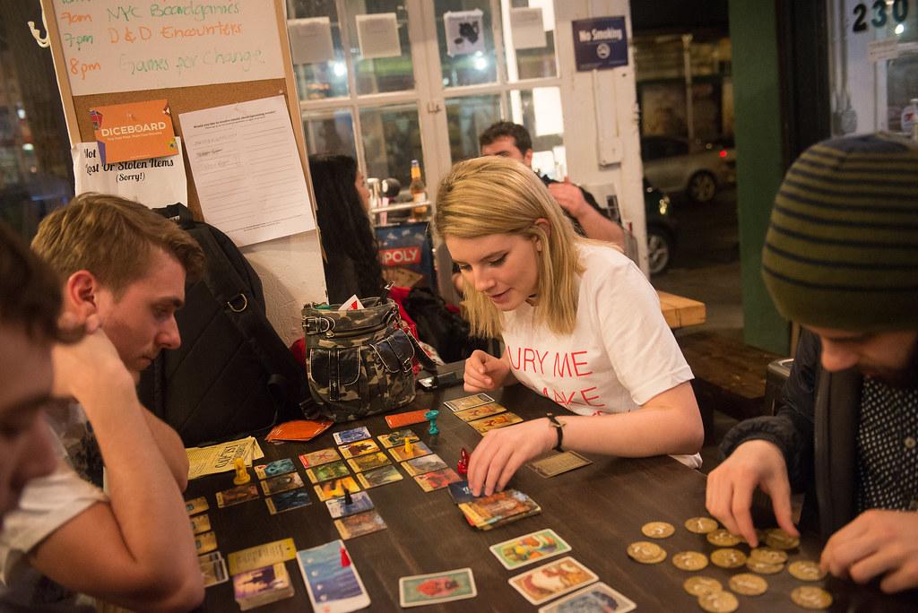 Several adults sit at a table playing a game with cards, tokens and other pieces.