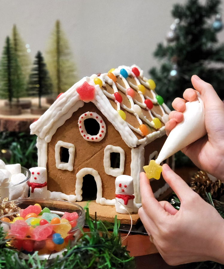 A person uses white icing to attach candies to a decorated gingerbread house.