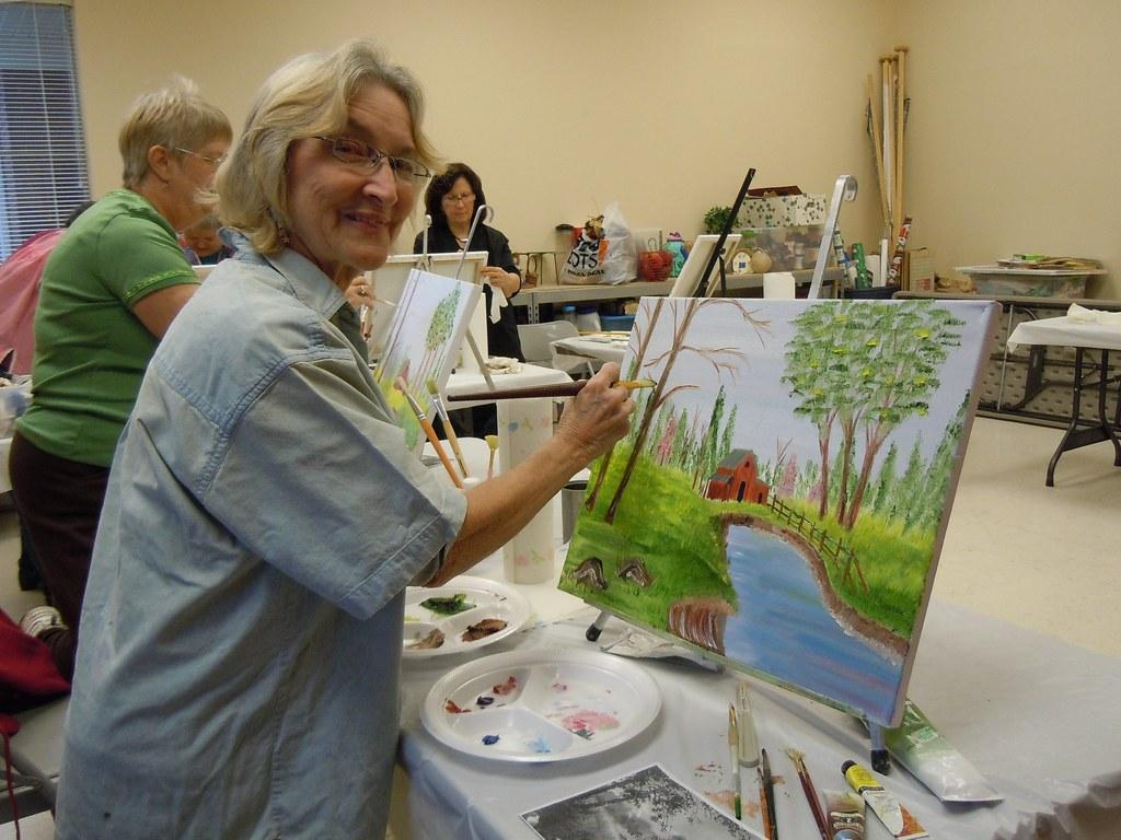 A senior woman paints a landscape featuring a river and barn.