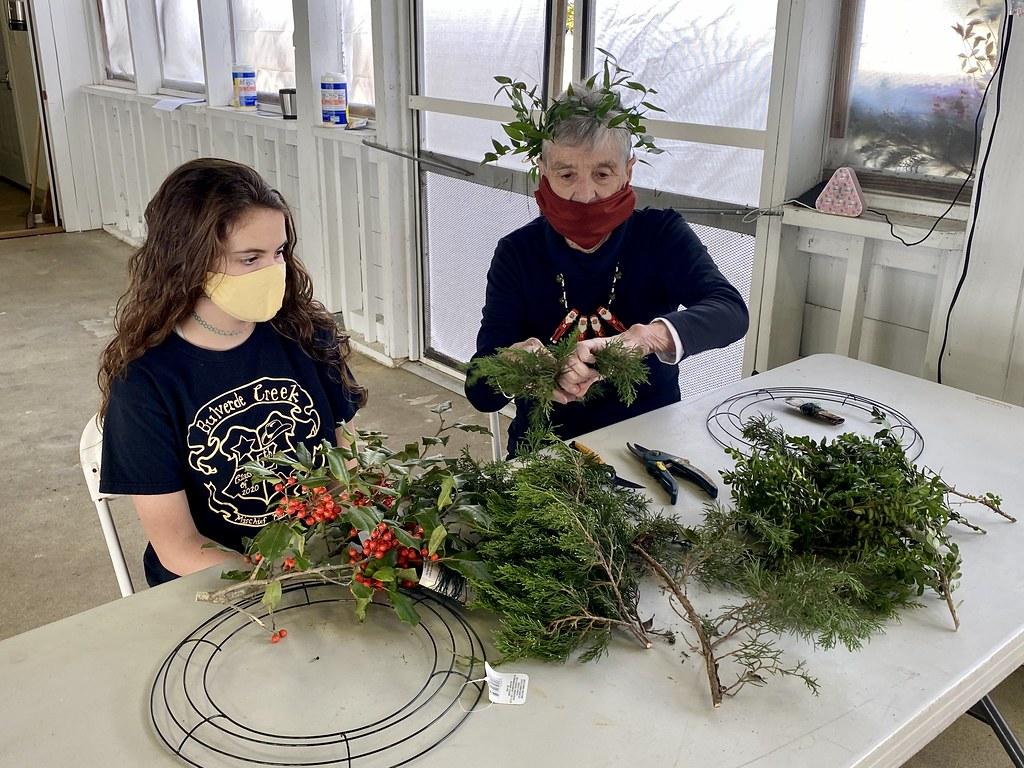 Two adults sit at a table, adding fronds to empty wreath forms.